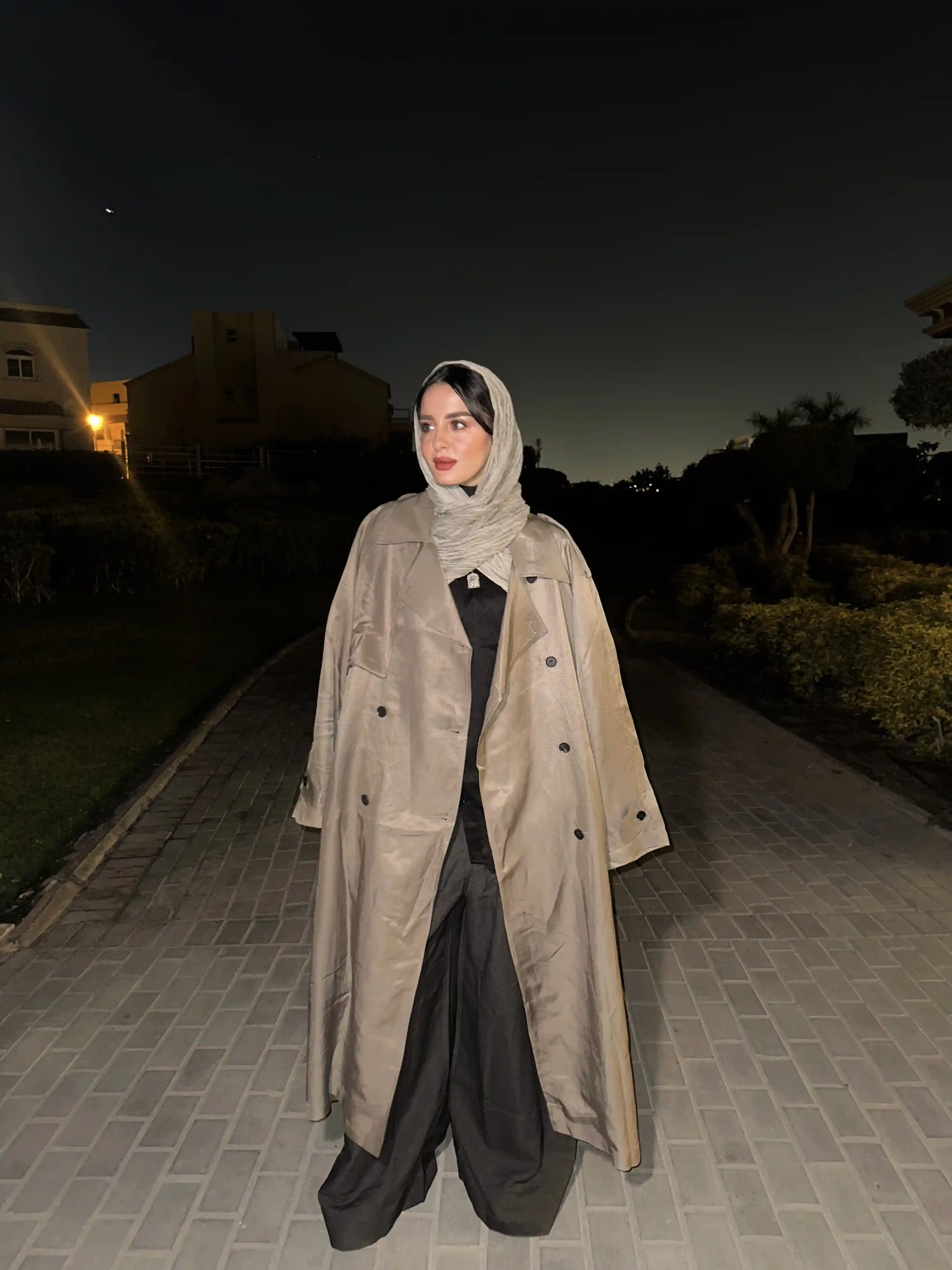 Person wearing a long beige coat and headscarf standing on a pathway at night.